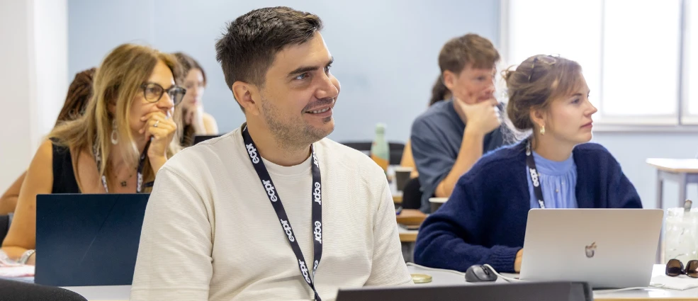 A group of people seated in a classroom setting, with a man in the foreground wearing an ECPR lanyard, smiling and looking to the side.